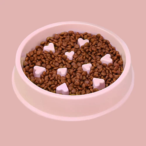 Pink slow feeder dog bowl with raised heart-shaped sections filled with dry dog food, shown on a plain white background.