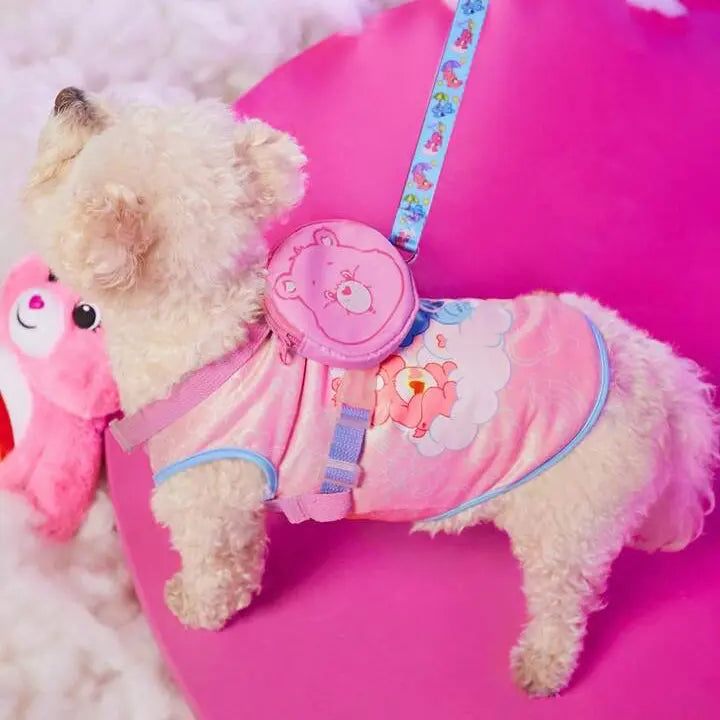 Small fluffy dog wearing a pink harness with a Care Bears design, matching leash, and a pink bear-shaped backpack, sitting on a pink surface.