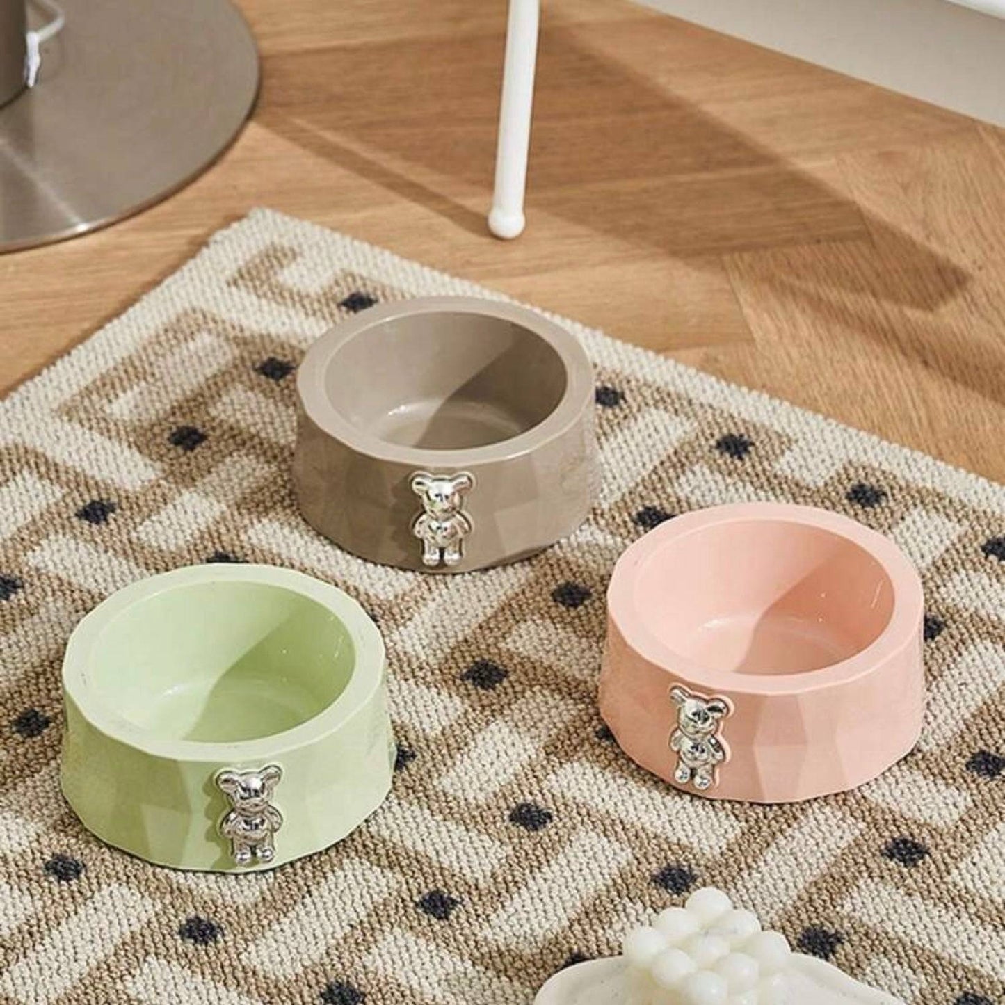 Three geometric dog bowls in lime green, grey, and pink, each with a metallic teddy bear decoration, arranged on a patterned rug.