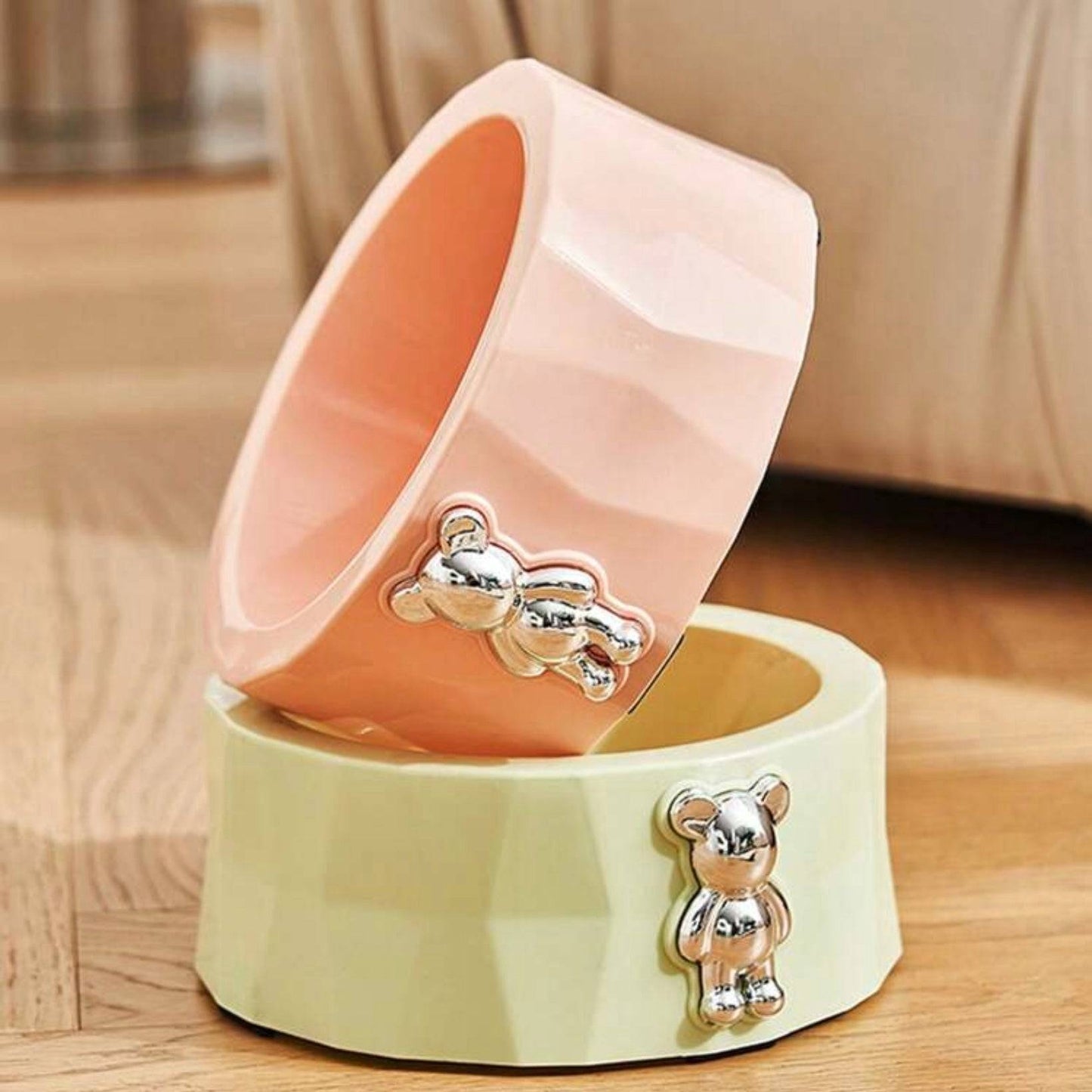 Two geometric dog bowls, one pink and one lime green, each with a metallic teddy bear decoration, stacked on a wooden floor.