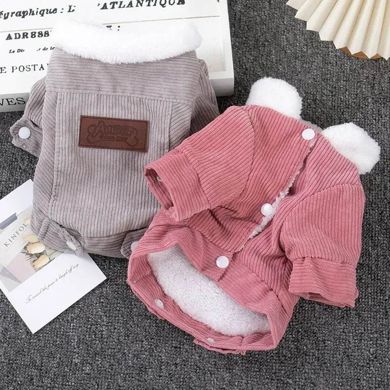 Two corduroy dog jackets, one in pink and one in grey, with white fleece lining and button details, displayed on a grey surface.