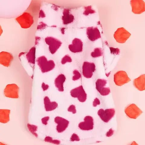 Flat lay of a pink heart print faux fur dog jacket, displayed on a pink background with scattered rose petals.