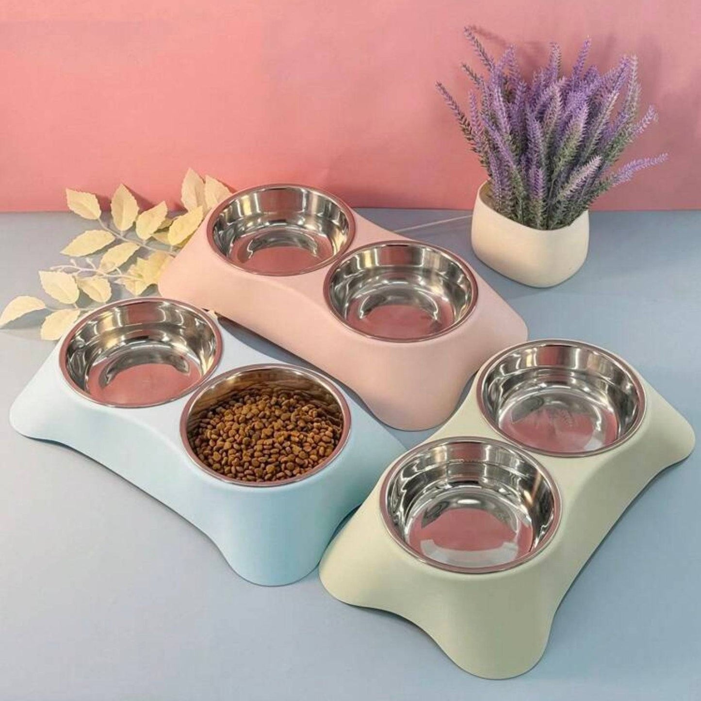 Three dual stainless steel dog bowl sets in lime, blue, and pink bone-shaped stands, arranged together with dry pet food in one bowl and decorative plants in the background.