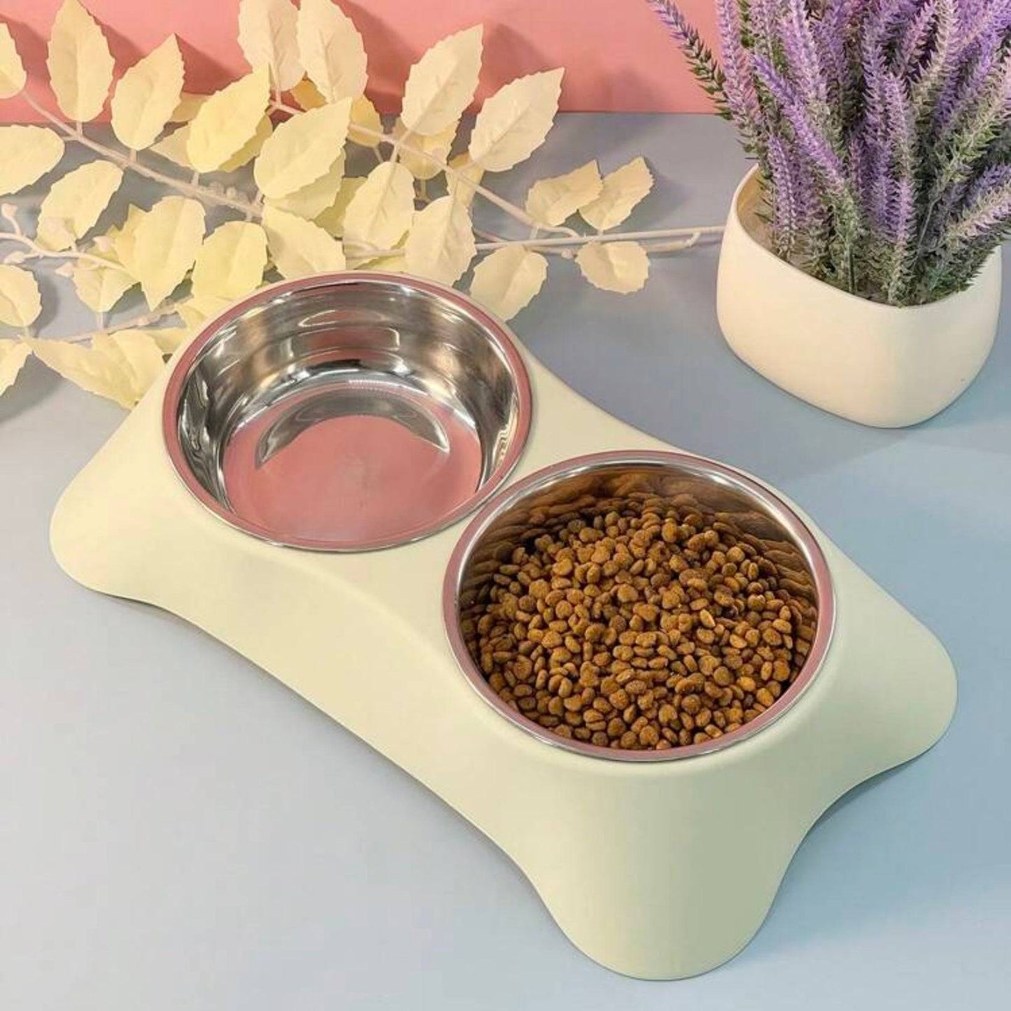 Dual stainless steel dog bowls on a lime green bone-shaped stand, with dry pet food in one bowl and decorative plants in the background.