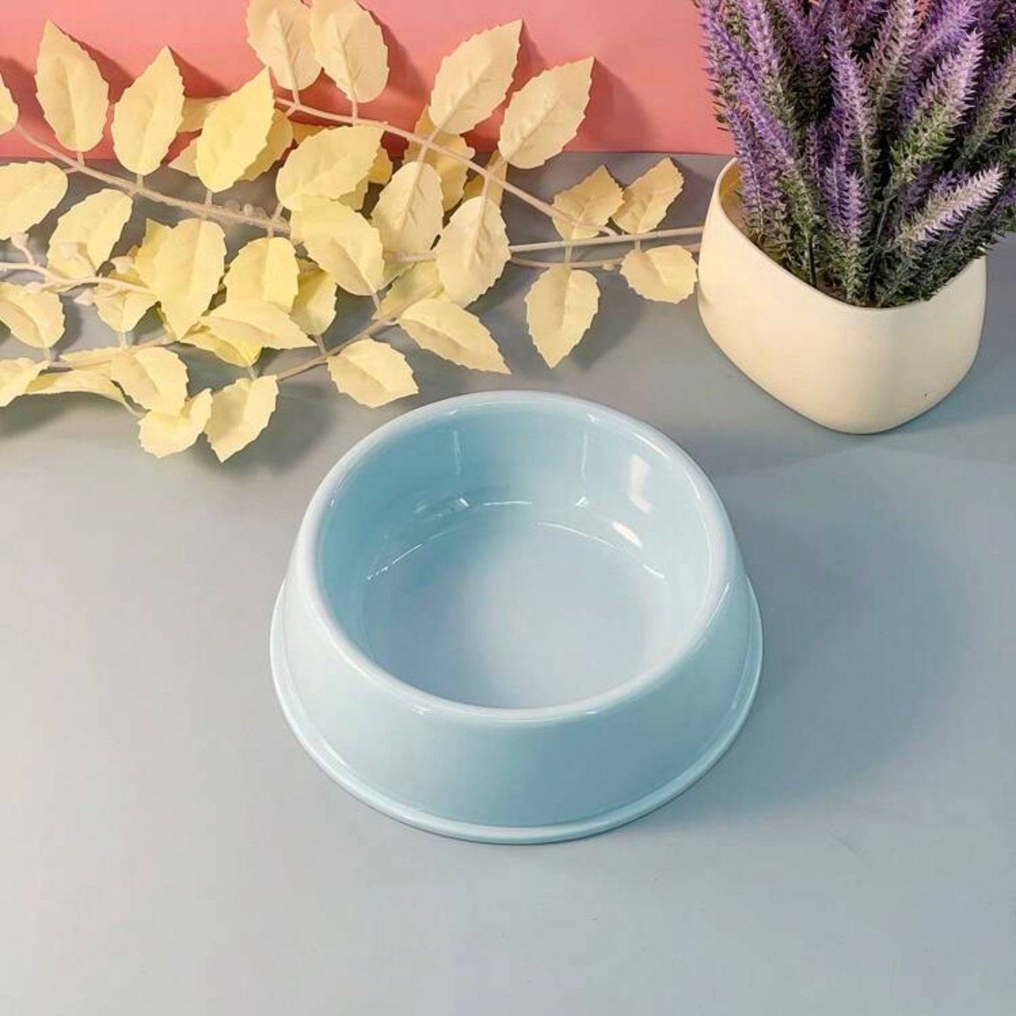 Pastel blue plastic dog bowl on a gray surface, with yellow artificial leaves and a potted purple plant in the background.