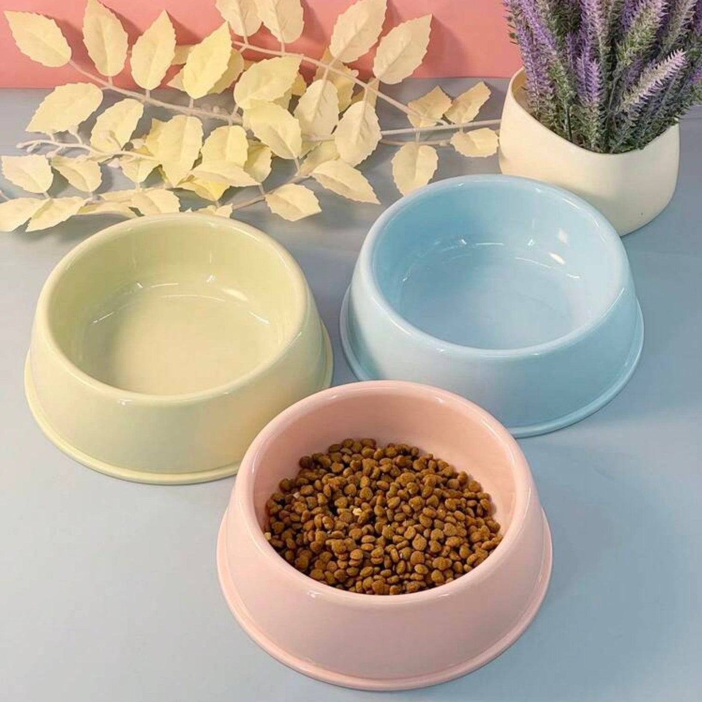 Three plastic dog bowls in lime green, blue, and pink (the pink bowl filled with kibble), arranged on a gray surface with yellow artificial leaves and a potted purple plant in the background.