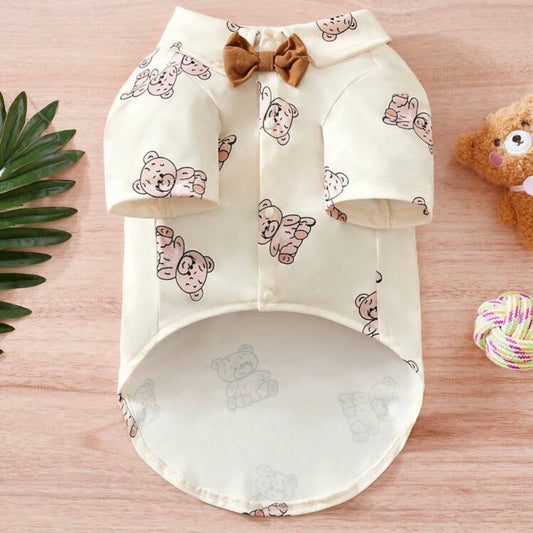 Front view of a cream-colored dog shirt with short sleeves and a brown bow tie, decorated with a teddy bear print pattern, laid out on a wooden surface.