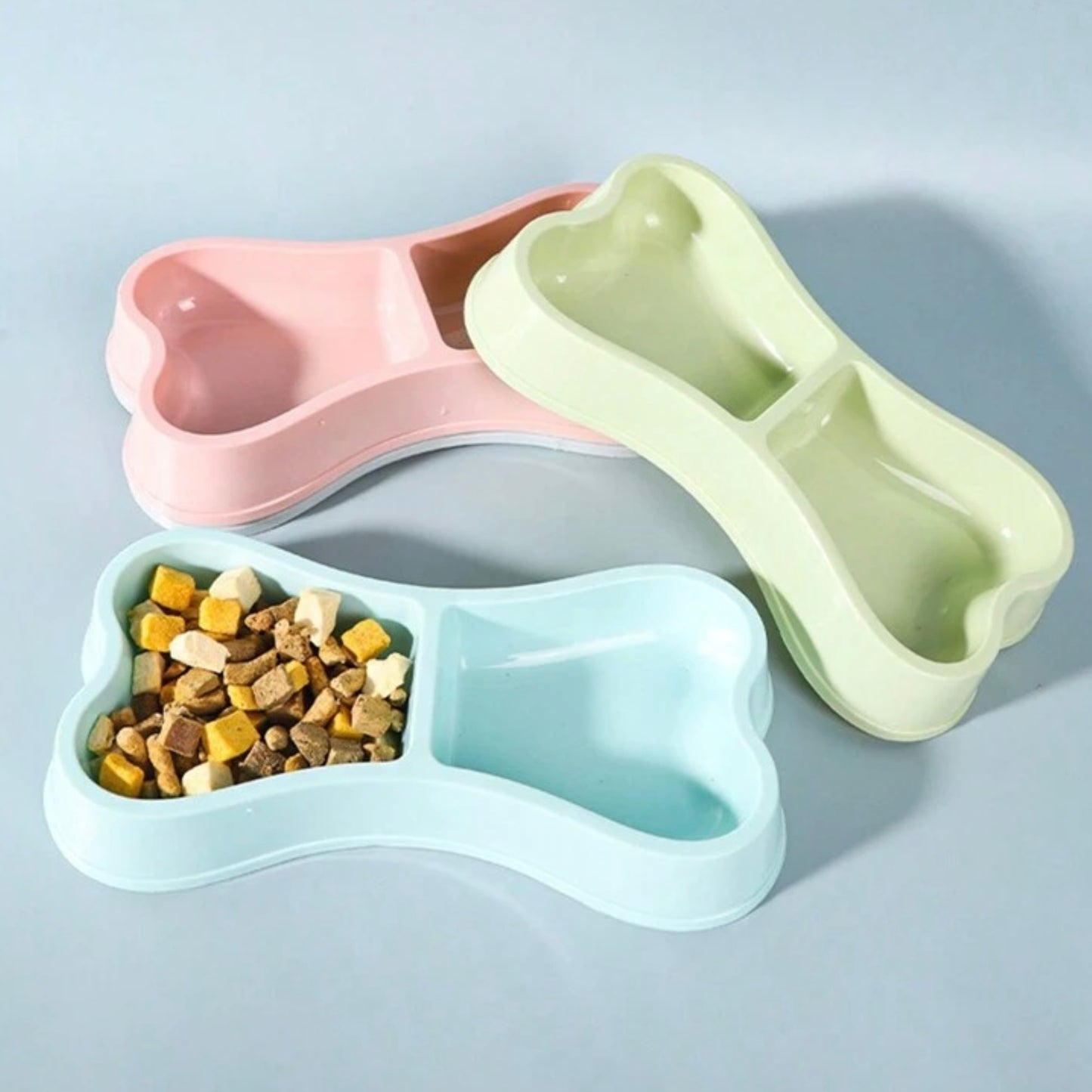 Three pastel-colored, bone-shaped double dog bowls in pink, green, and blue, with one bowl filled with dog food.