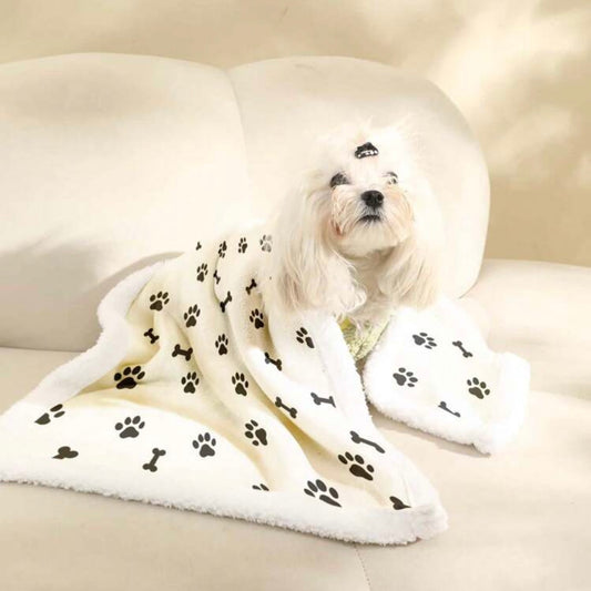 Small white dog sitting on a beige sofa, wrapped in a white dog blanket with black paw prints and bone patterns.