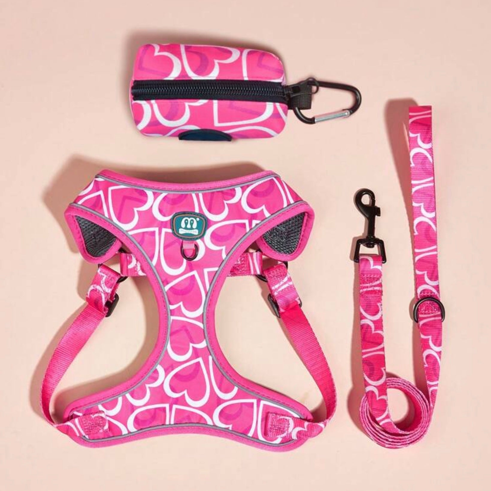 Set of matching pink dog accessories with a heart pattern, including a harness, leash, and poop bag holder, arranged on a light background.