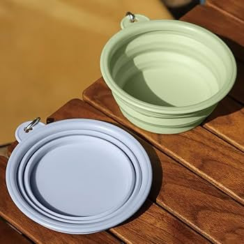 Collapsible silicone dog bowls in green and blue, one expanded and one folded flat, with carabiner clips, placed on a wooden surface.