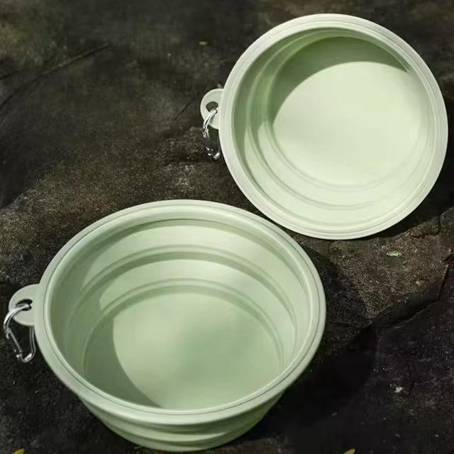 Two light green collapsible dog bowls, both expanded and displayed outdoors on a dark textured surface, each with a carabiner clip.