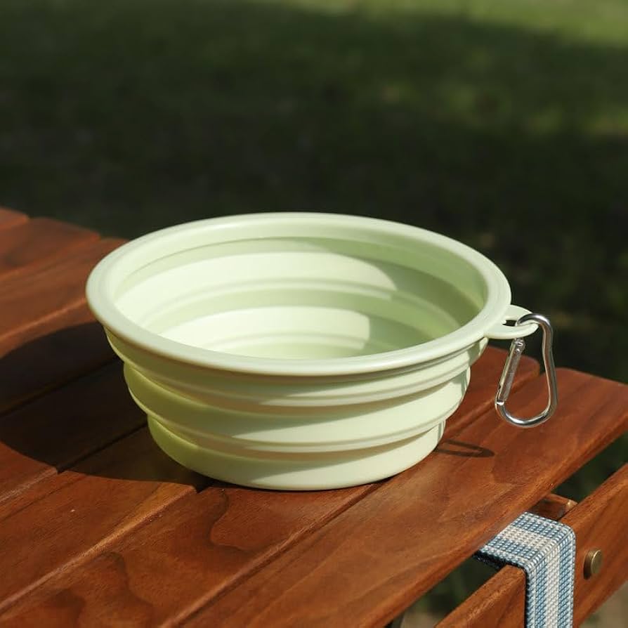Light green collapsible dog bowl fully expanded with a carabiner clip, sitting on a wooden outdoor bench.