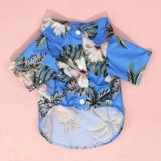 Blue Hawaiian-style dog shirt with white buttons and a tropical hibiscus floral, palm leaf and pineapple print, shown from the front on a pink background.