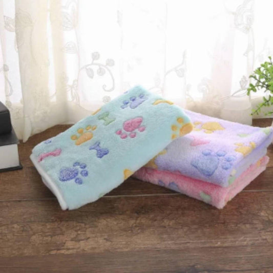 Three folded blankets, one mint, one lilac and one pink, all decorated with colorful paw prints and bone patterns, placed on a wooden surface.