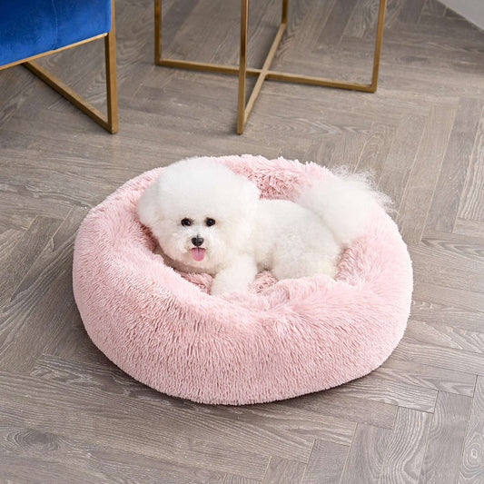 A fluffy white Bichon Frise lying comfortably in a round, pink, plush dog bed on a light wood floor.