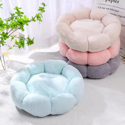 Stack of three plush round dog beds in cream, pink, and gray, with a light blue bed in front, placed in a bright room with a plant nearby.