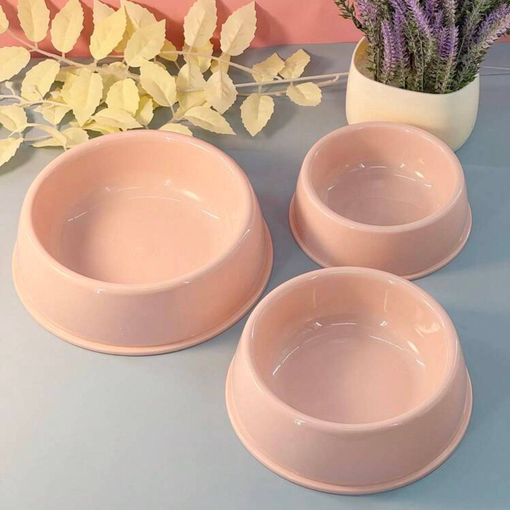 Three pastel pink ceramic dog bowls of different sizes on a gray surface, with yellow artificial leaves and a potted purple plant in the background.